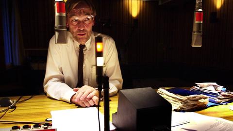 Wir sehen einen Mann, der in einem Radiostudio sitzt und einen Text einsprechen möchte. 