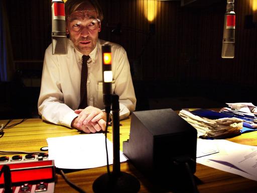 Wir sehen einen Mann, der in einem Radiostudio sitzt und einen Text einsprechen möchte. 