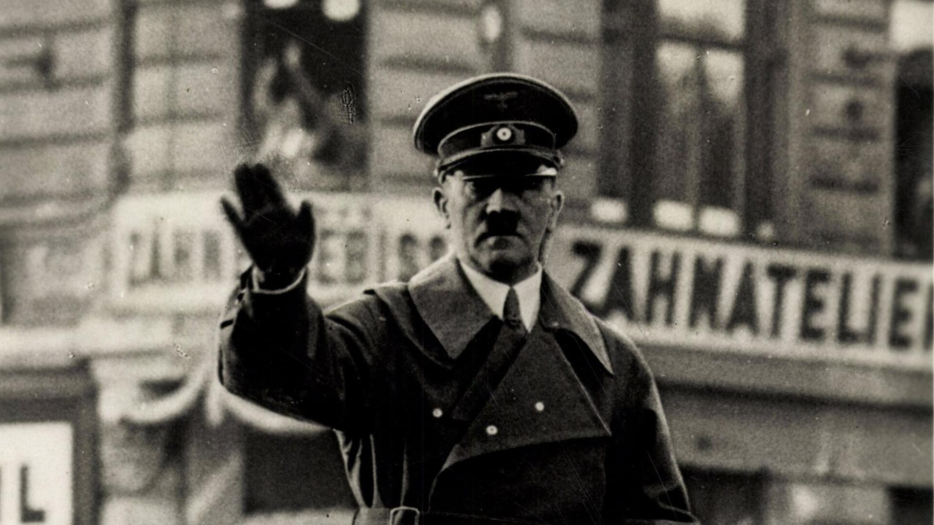 Adolf Hitler steht in einem fahrenden Auto und winkt.