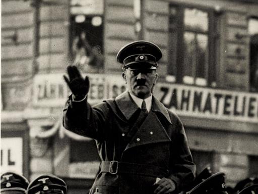 Adolf Hitler steht in einem fahrenden Auto und winkt.