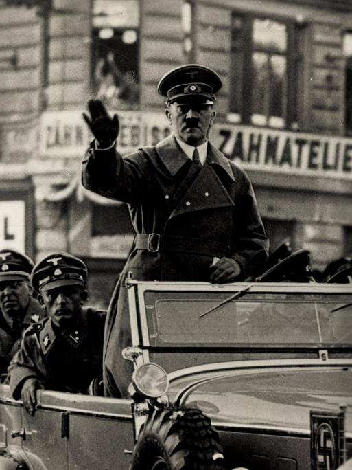 Adolf Hitler steht in einem fahrenden Auto und winkt.