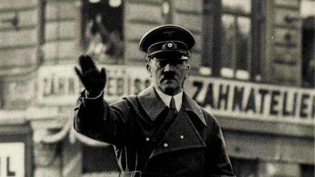 Adolf Hitler steht in einem fahrenden Auto und winkt.