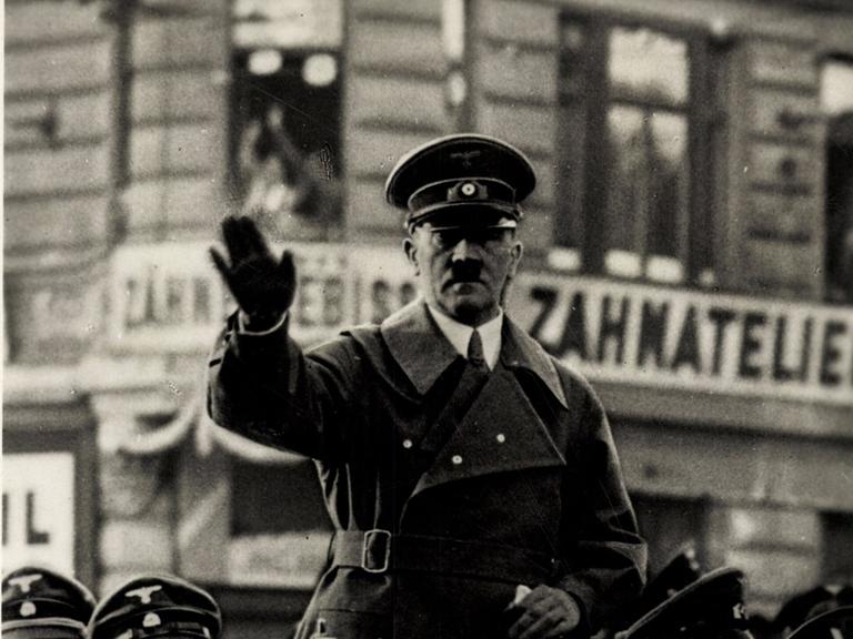Adolf Hitler steht in einem fahrenden Auto und winkt.