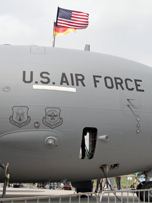 Das Foto zeigt ein graues Transport-Militärflugzeug am Boden. Das Flugzeug trägt die schwarze Aufschrift „U.S. Air Force“. Auf dem Flugzeug sind die Flaggen der USA und Deutschlands angebracht. 