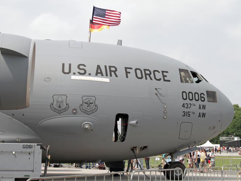 Das Foto zeigt ein graues Transport-Militärflugzeug am Boden. Das Flugzeug trägt die schwarze Aufschrift „U.S. Air Force“. Auf dem Flugzeug sind die Flaggen der USA und Deutschlands angebracht. 