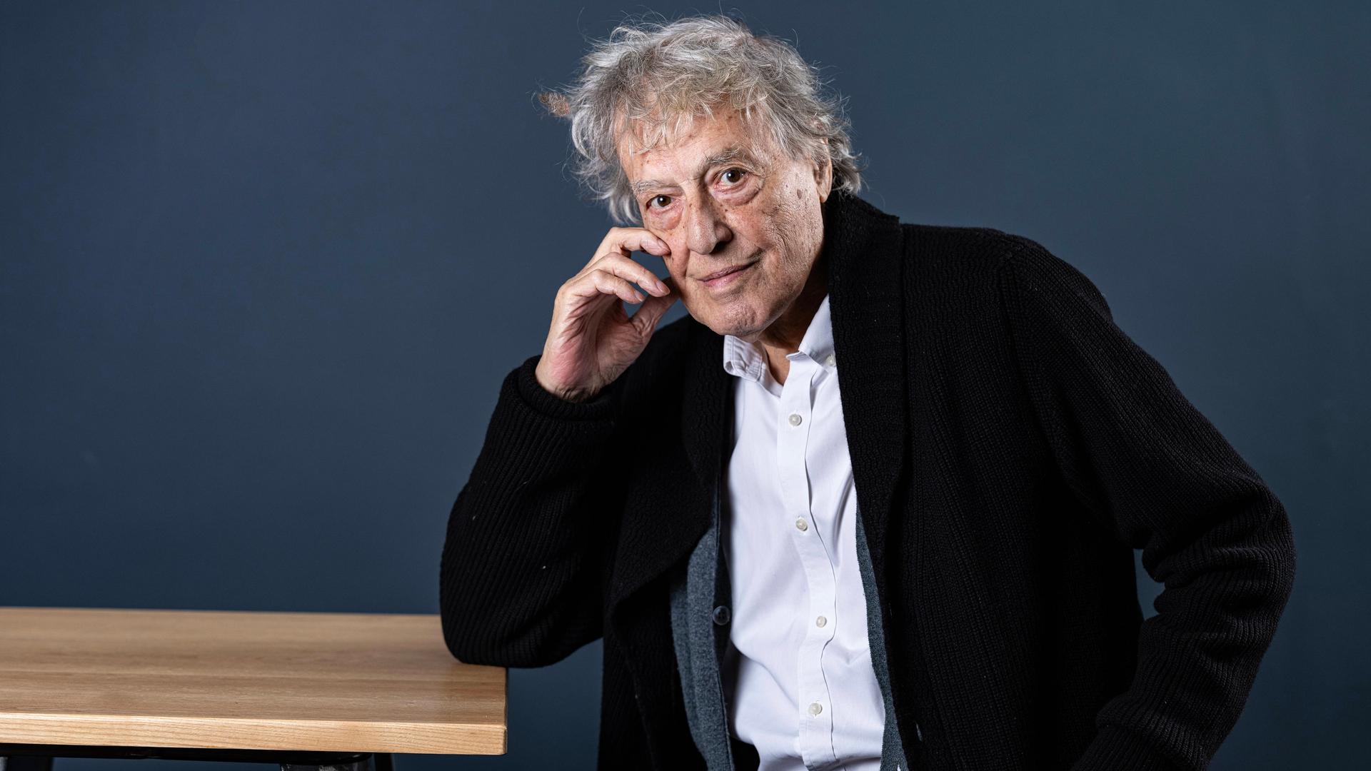 Der britische Dramatiker Tom Stoppard sitzt seitlich an einem Tisch, stützt den rechten Ellenbogen auf die Tischkante und fast sich mit der rechten Hand an die rechte Wange