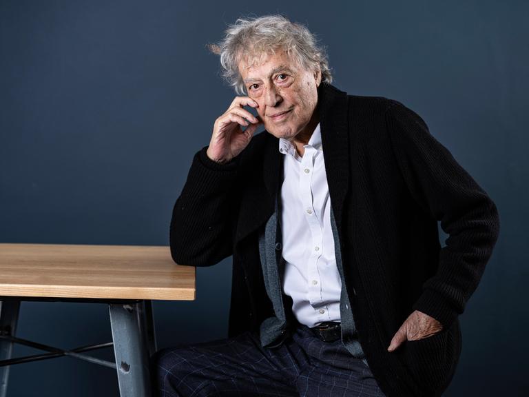 Der britische Dramatiker Tom Stoppard sitzt seitlich an einem Tisch, stützt den rechten Ellenbogen auf die Tischkante und fast sich mit der rechten Hand an die rechte Wange Der britische Dramatiker Tom Stoppard sitzt seitlich an einem Tisch, stützt den rechten Ellenbogen auf die Tischkante und fast sich mit der rechten Hand an die rechte Wange