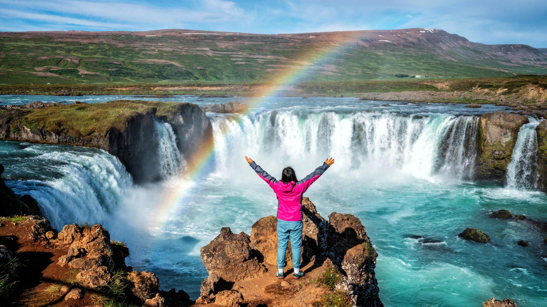 Eine Person in der Rückansicht blickt mit erhobenen Armen auf den Godafoss (isländisch: Wasserfall der Götter), einen berühmten Wasserfall in Island vor atemberaubener Landschaft, über die sich ein Regenbogen spannt. Eine Person in der Rückansicht blickt mit erhobenen Armen auf den Godafoss (isländisch: Wasserfall der Götter), einen berühmten Wasserfall in Island vor atemberaubener Landschaft, über die sich ein Regenbogen spannt.