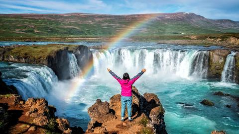 Eine Person in der Rückansicht blickt mit erhobenen Armen auf den Godafoss (isländisch: Wasserfall der Götter), einen berühmten Wasserfall in Island vor atemberaubener Landschaft, über die sich ein Regenbogen spannt.