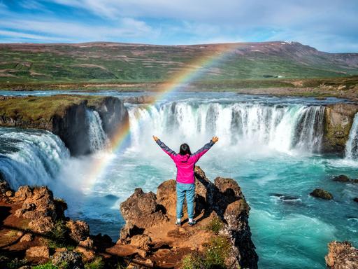 Eine Person in der Rückansicht blickt mit erhobenen Armen auf den Godafoss (isländisch: Wasserfall der Götter), einen berühmten Wasserfall in Island vor atemberaubener Landschaft, über die sich ein Regenbogen spannt.