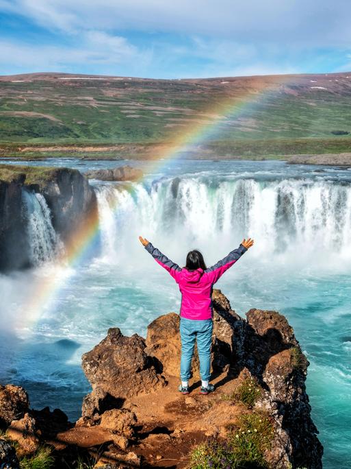 Eine Person in der Rückansicht blickt mit erhobenen Armen auf den Godafoss (isländisch: Wasserfall der Götter), einen berühmten Wasserfall in Island vor atemberaubener Landschaft, über die sich ein Regenbogen spannt. Eine Person in der Rückansicht blickt mit erhobenen Armen auf den Godafoss (isländisch: Wasserfall der Götter), einen berühmten Wasserfall in Island vor atemberaubener Landschaft, über die sich ein Regenbogen spannt.