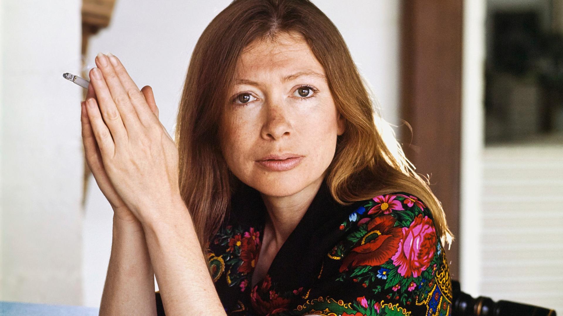 Kopf- und Schulterporträt der Schriftstellerin Joan Didion in ihrem Haus in Malibu, Kalifornien; sie sitzt in einem Stuhl, stützt die Ellbogen auf den Tisch vor ihr, legt die Hände mit den Handflächen aneinander und hält eine Zigarette.  Sie trägt einen schwarzen Schal mit bunten Blumen und Mustern, und ihr Haar hängt ihr bis zu den Schultern herab.