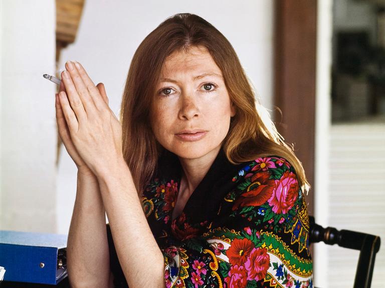 Kopf- und Schulterporträt der Schriftstellerin Joan Didion in ihrem Haus in Malibu, Kalifornien; sie sitzt in einem Stuhl, stützt die Ellbogen auf den Tisch vor ihr, legt die Hände mit den Handflächen aneinander und hält eine Zigarette.  Sie trägt einen schwarzen Schal mit bunten Blumen und Mustern, und ihr Haar hängt ihr bis zu den Schultern herab.