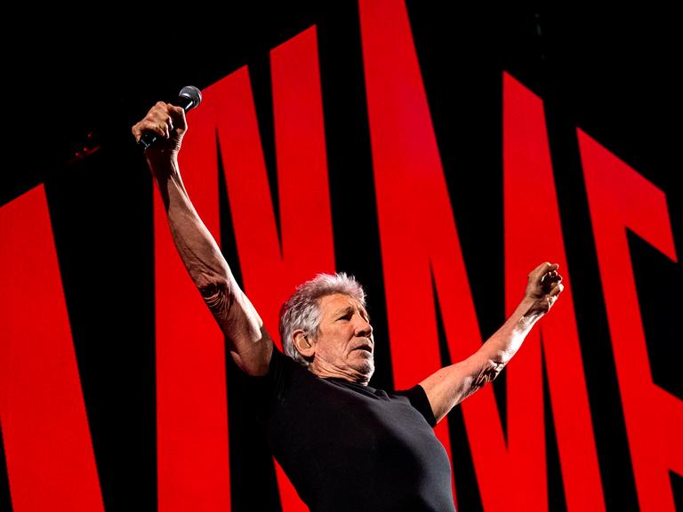Roger Waters auf der Bühne in Hamburg mit ausgebreiteten Armen. Hinter ihm rote Symbole vor schwarzem Hintergrund.