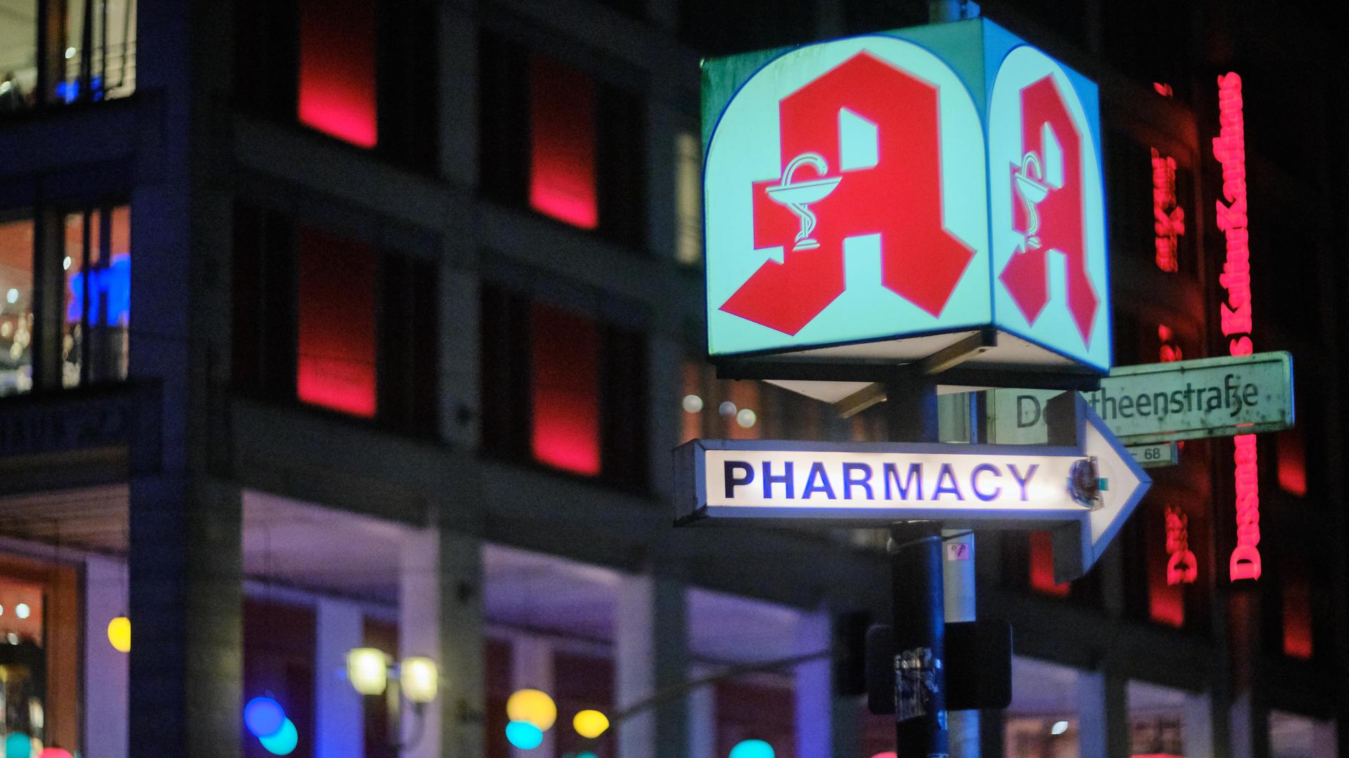 Eine Leuchtreklame mit einem Apothekensymbol und einem beleuchteten Pfeil mit der Aufschrift "Pharmacy" leuchtet in der Nacht an einer Berliner Kreuzung. 