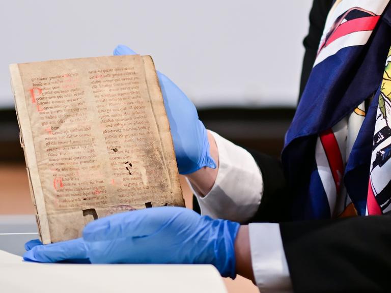 Zu sehen ist eine alte Handschrift. Ein Person mit blauen Handschuhen hält das Dokument fest.