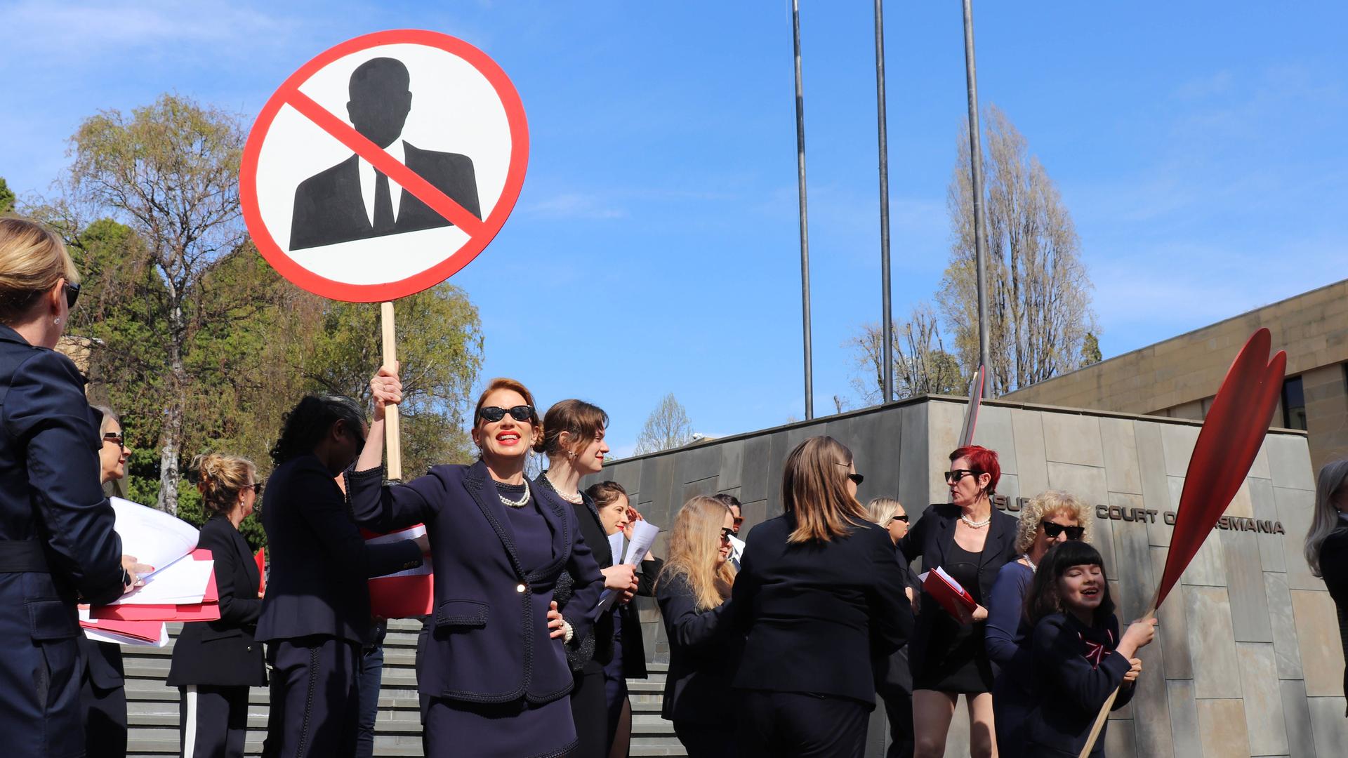 Frauen in schwarzen Anziehsachen stehen vor einem Gerichtsgebäude. Eine trägt ein rundes Schild, auf dem ein stilisierter Mann zu sehen ist. Er ist durchgestrichen.