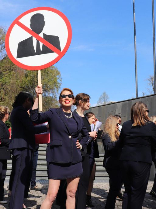 Frauen in schwarzen Anziehsachen stehen vor einem Gerichtsgebäude. Eine trägt ein rundes Schild, auf dem ein stilisierter Mann zu sehen ist. Er ist durchgestrichen.