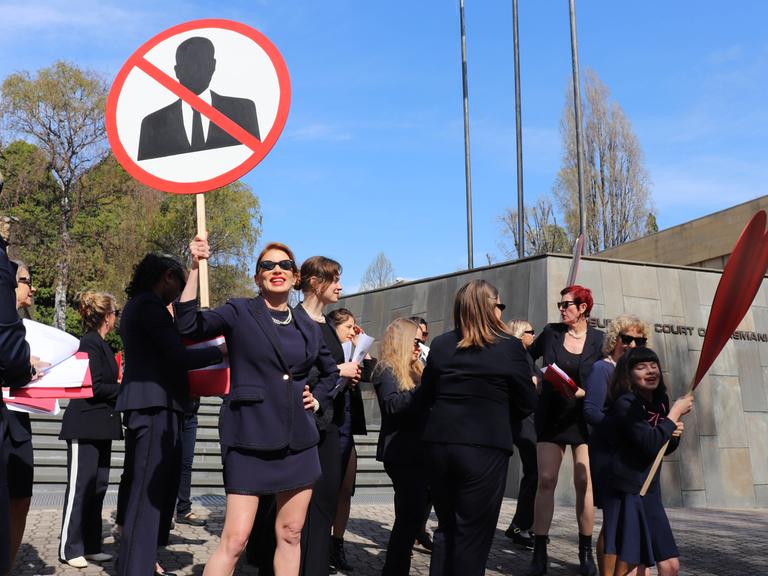 Frauen in schwarzen Anziehsachen stehen vor einem Gerichtsgebäude. Eine trägt ein rundes Schild, auf dem ein stilisierter Mann zu sehen ist. Er ist durchgestrichen.