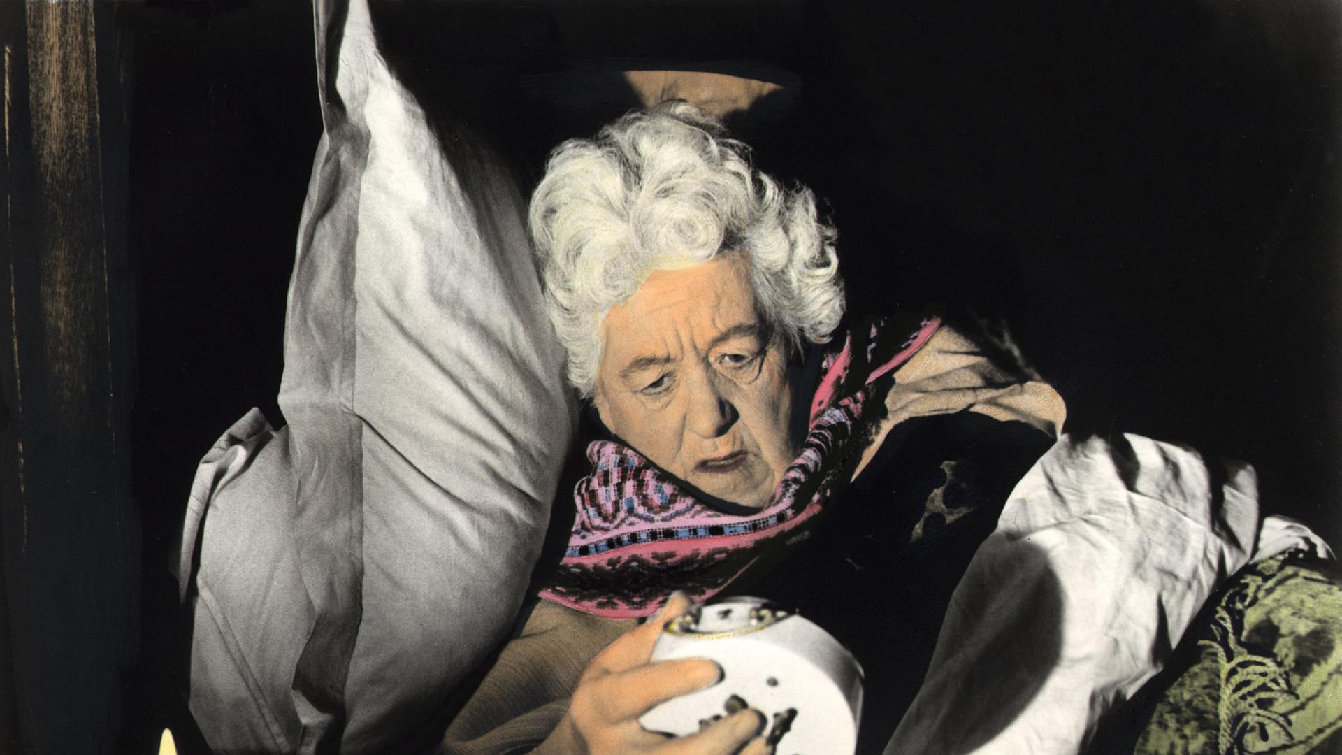 Ein koloriertes Film-Still zeigt die Schauspielerin Margaret Rutherford  in der Rolle der ergrauten Miss Marple schlafend, einen Wecker umarmend