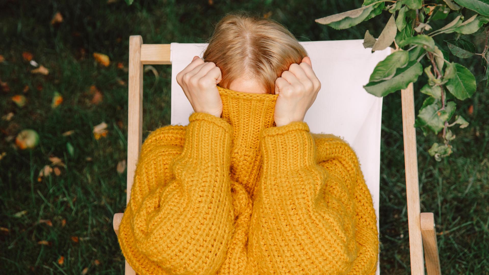 Eine Frau in gelbem Pullover versteckt ihr Gesicht im hochgezogenen Pulloverkragen.
