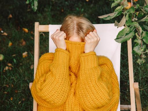 Eine Frau in gelbem Pullover versteckt ihr Gesicht im hochgezogenen Pulloverkragen.