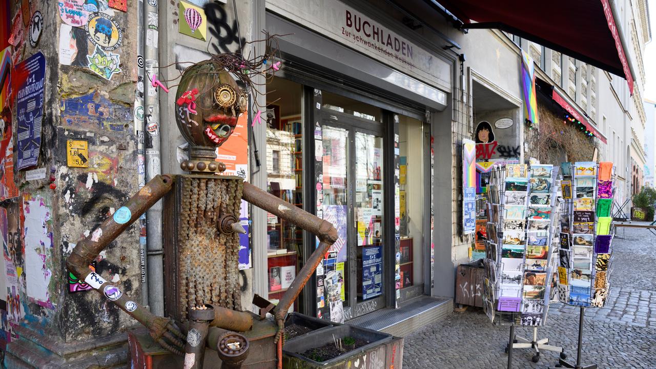 Fassade des Buchladens „Zur schwankenden Weltkugel“ in Berlin, davor eine mit antifaschistischen Stickern beklebte Blechskulptur neben Blumenkästen und Postkartenständern.
