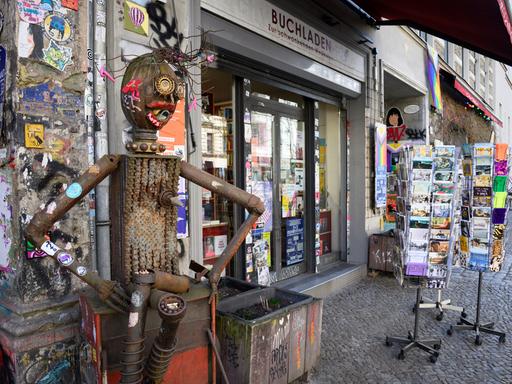 Fassade des Buchladens „Zur schwankenden Weltkugel“ in Berlin, davor eine mit antifaschistischen Stickern beklebte Blechskulptur neben Blumenkästen und Postkartenständern.