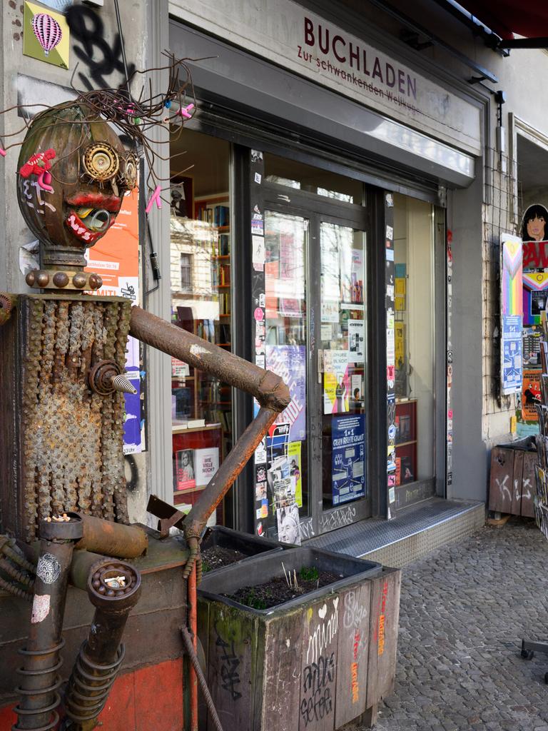 Fassade des Buchladens „Zur schwankenden Weltkugel“ in Berlin, davor eine mit antifaschistischen Stickern beklebte Blechskulptur neben Blumenkästen und Postkartenständern.