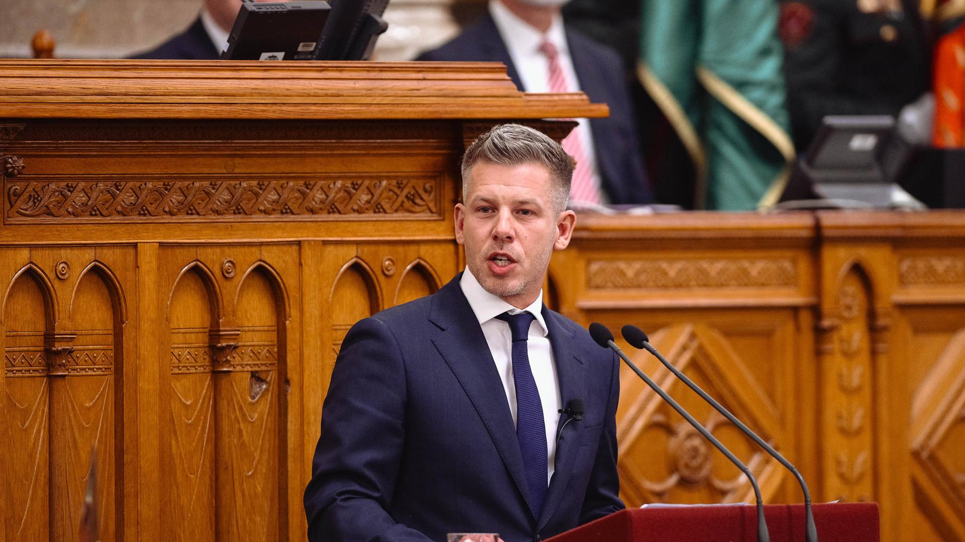 Peter Magyar steht vor einer Wand aus Holz an einem Redner-Pult. Er trägt einen Anzug mit Krawatte und spricht in ein Mikrofon.