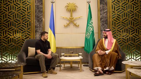 Der ukrainische Präsident Selenskyj und der saudische Kronprinz Mohammed bin Salman sitzen nebeneinander und führen ein Gespräch. Im Hintergrund sind die Flaggen der Ukraine und von Saudi-Arabien.