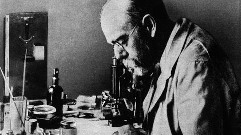 Schwarz-Weiß-Aufnahme: Robert Koch blickt in ein Mikroskop. 