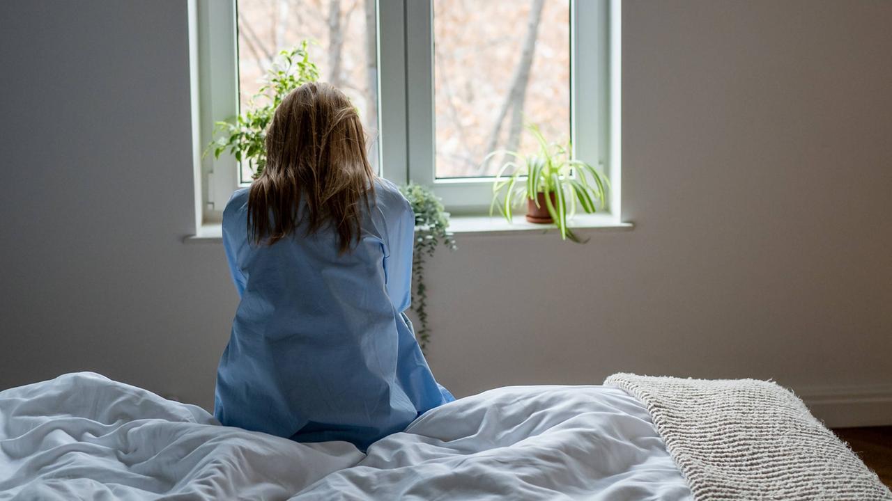 Frau sitzt auf einem weißen Bett und schaut aus dem Fenster. Sie hat lange blonde Haare und eine blaue Bluse an. 