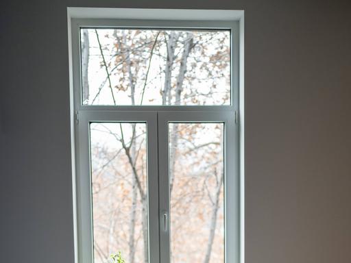 Frau sitzt auf einem weißen Bett und schaut aus dem Fenster. Sie hat lange blonde Haare und eine blaue Bluse an. 