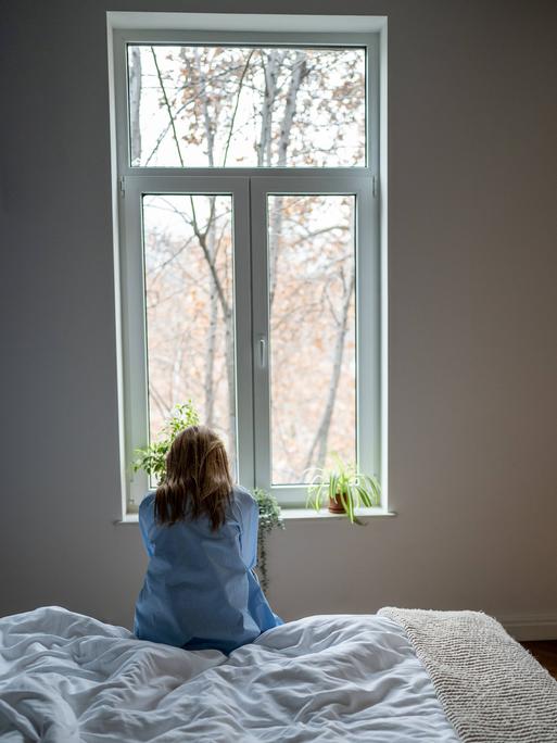 Frau sitzt auf einem weißen Bett und schaut aus dem Fenster. Sie hat lange blonde Haare und eine blaue Bluse an. 