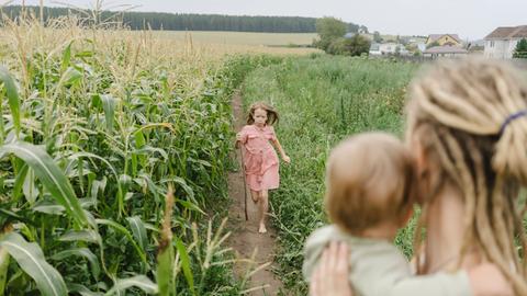 Ein Mädchen läuft auf einem Maisfeld seiner Mutter entgegen, im Hintergrund ist ein Dorf zu sehen.