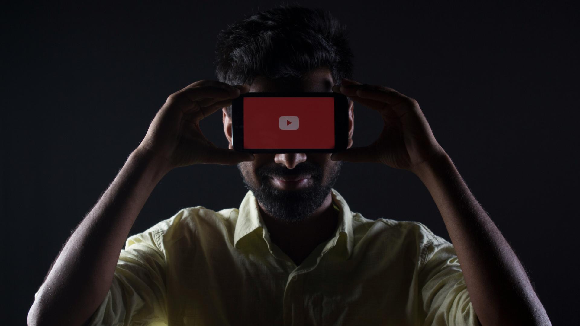 Ein dunkelhaariger Mann mit hellem Hemd hält sich in einem dunklen Raum ein Handy wie eine Maske vor die Augen, auf dem ein roter Playbutton zu sehen ist. 