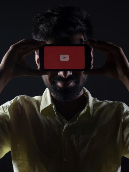 Ein dunkelhaariger Mann mit hellem Hemd hält sich in einem dunklen Raum ein Handy wie eine Maske vor die Augen, auf dem ein roter Playbutton zu sehen ist. 