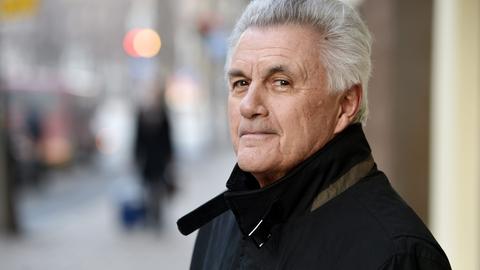 John Irving steht im Freien und blickt freundlich lächelnd in die Kamera. Der Hintergrund ist unscharf.