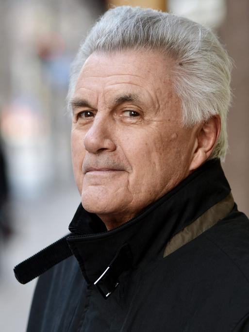 John Irving steht im Freien und blickt freundlich lächelnd in die Kamera. Der Hintergrund ist unscharf.