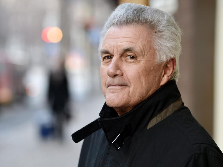 John Irving steht im Freien und blickt freundlich lächelnd in die Kamera. Der Hintergrund ist unscharf.