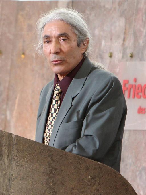 Boualem Sansal steht auf einem Podium und spricht ins Mikrofon. Er trägt das lange graue Haar zu einem Zopf gebunden und ein graues Sakko.