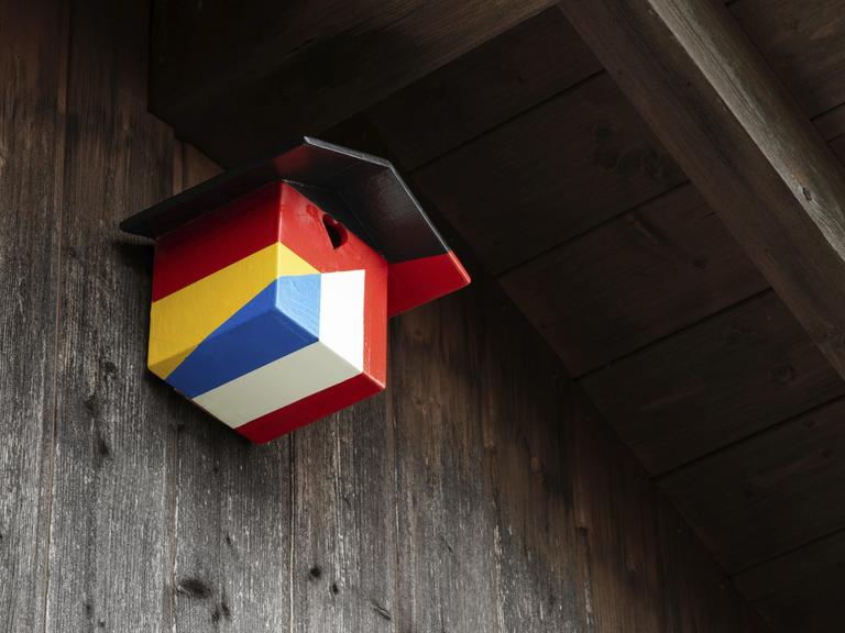 Ein Nistkasten für Vögel in den Farben der deutschen und französischen Nationalflagge hängt an einer hölzernen Hauswand. Ein Nistkasten für Vögel in den Farben der deutschen und französischen Nationalflagge hängt an einer hölzernen Hauswand.