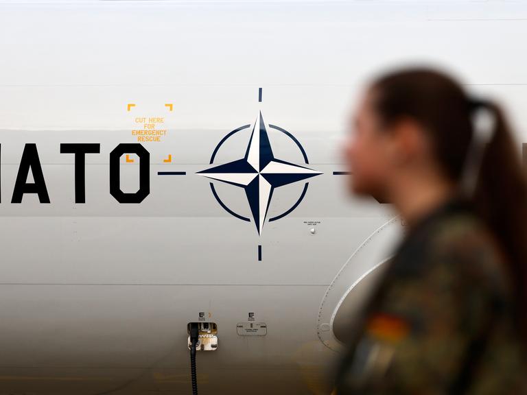 Eine Soldatin steht vor einem NATO-Logo. Das Logo ist auf einem AWACS-Flugzeug angebracht. Eine Soldatin steht vor einem NATO-Logo. Das Logo ist auf einem AWACS-Flugzeug angebracht.