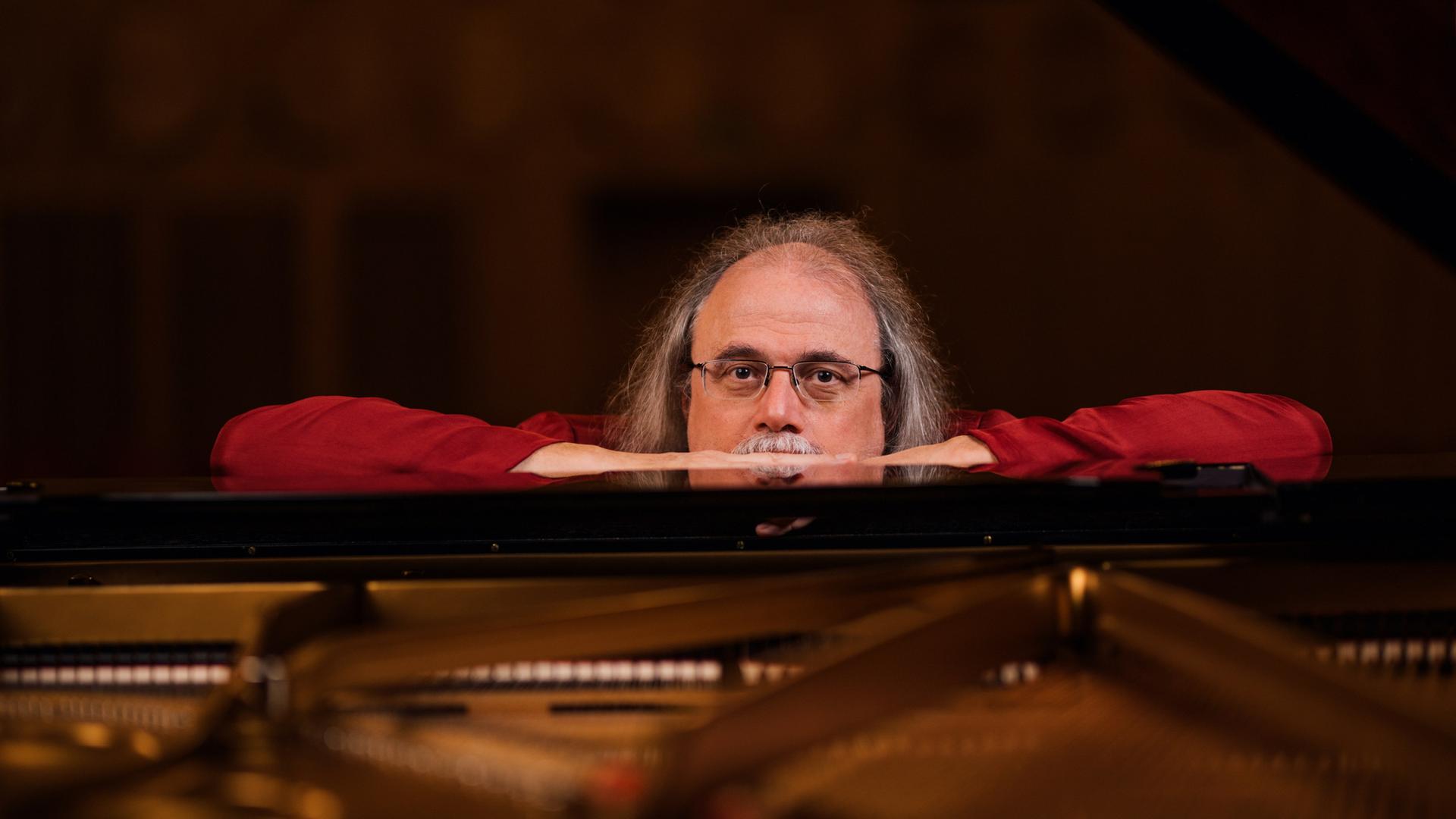 Ein Mann mit Brille und rotem Shirt lehnt dem Deckel eines Konzertflügels und schaut knapp über dem Instrument in die Kamera