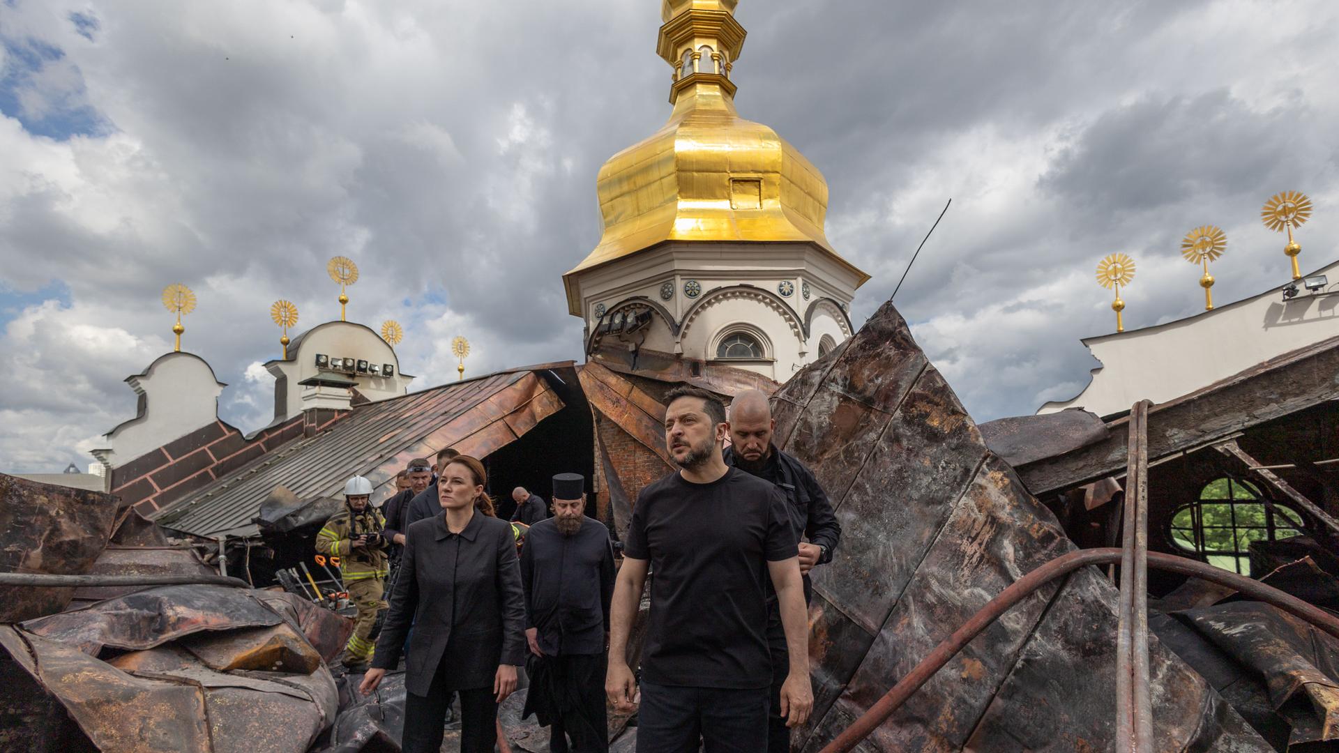 Der ukrainische Präsident Wolodymyr Selenskyj steht vor dem zerstörten Dachstuhl der Entschlafungs-Kathedrale. Man sieht, dass die Dachkonstruktion verbogen ist. Der ukrainische Präsident Wolodymyr Selenskyj steht vor dem zerstörten Dachstuhl der Entschlafungs-Kathedrale. Man sieht, dass die Dachkonstruktion verbogen ist.