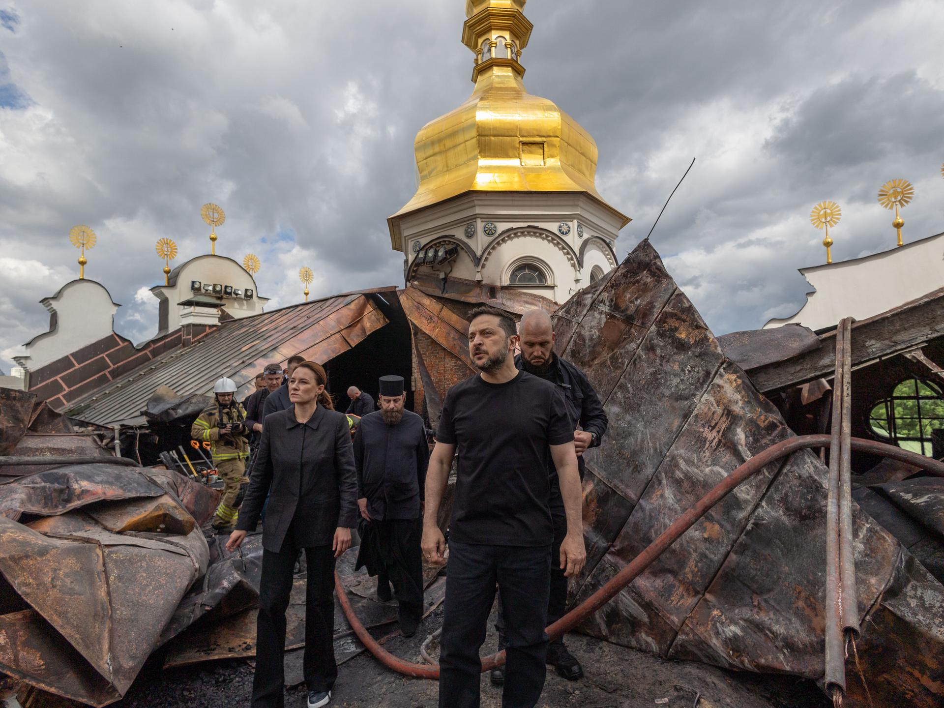 Der ukrainische Präsident Wolodymyr Selenskyj steht vor dem zerstörten Dachstuhl der Entschlafungs-Kathedrale. Man sieht, dass die Dachkonstruktion verbogen ist.