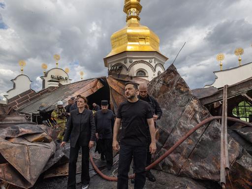 Der ukrainische Präsident Wolodymyr Selenskyj steht vor dem zerstörten Dachstuhl der Entschlafungs-Kathedrale. Man sieht, dass die Dachkonstruktion verbogen ist.