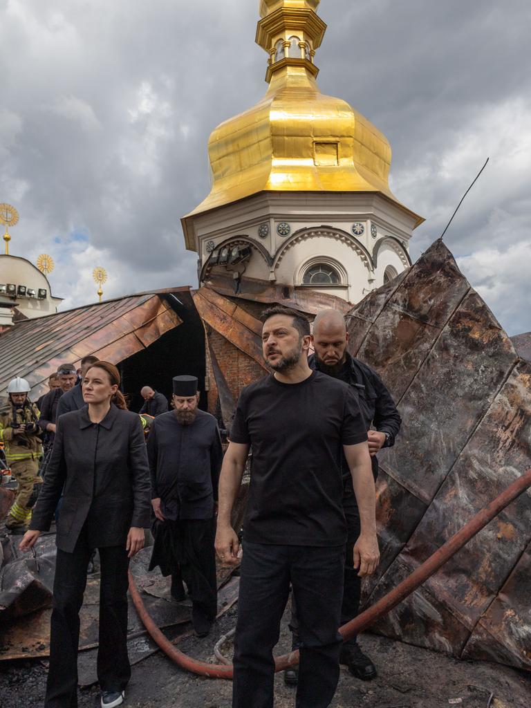 Der ukrainische Präsident Wolodymyr Selenskyj steht vor dem zerstörten Dachstuhl der Entschlafungs-Kathedrale. Man sieht, dass die Dachkonstruktion verbogen ist.