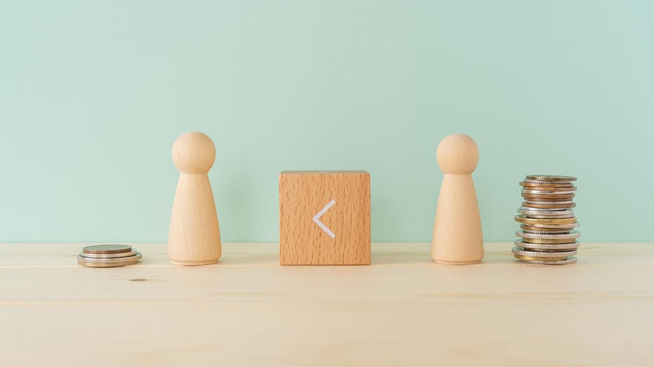 Zwei Spielfiguren aus Holz stehen auf einem Tisch. Dazwischen ein Holzklötzchen, auf dem das 
Größer-als-Zeichen zu sehen ist, es zeigt nach links. Neben der linken Figur stapeln sich vier Geldstücke, neben der rechten Figur deutlich mehr.
